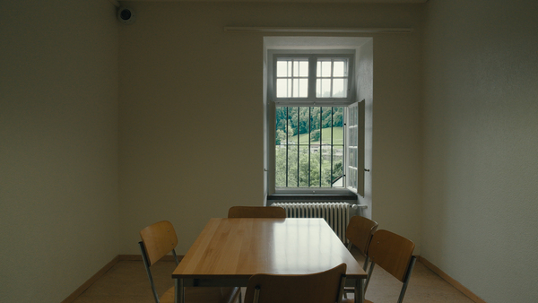 Filmstill aus Mitgefangen / Lomotion enger Raum mit vergittertem fenster am ende. drinnen steht ein tisch mit zwei stühlen
